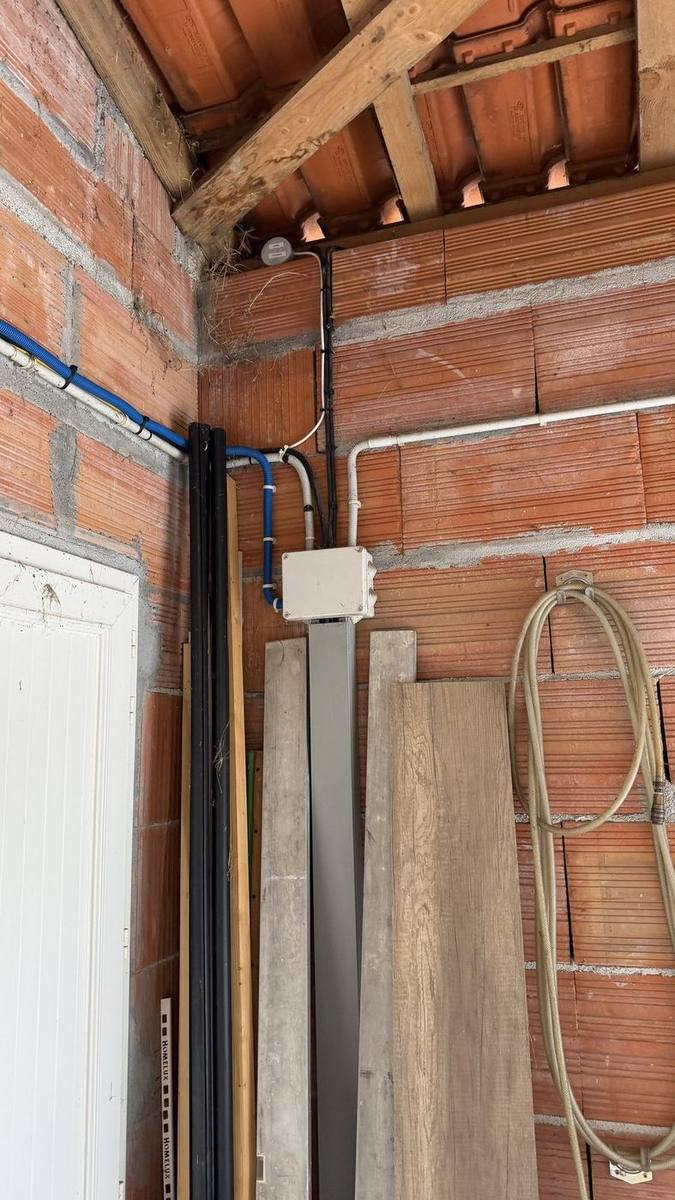 Boîtier de dérivation blanc fixé au mur de briques, où aboutissent la gaine ICTA bleue et les conduits blancs, sous la charpente du local