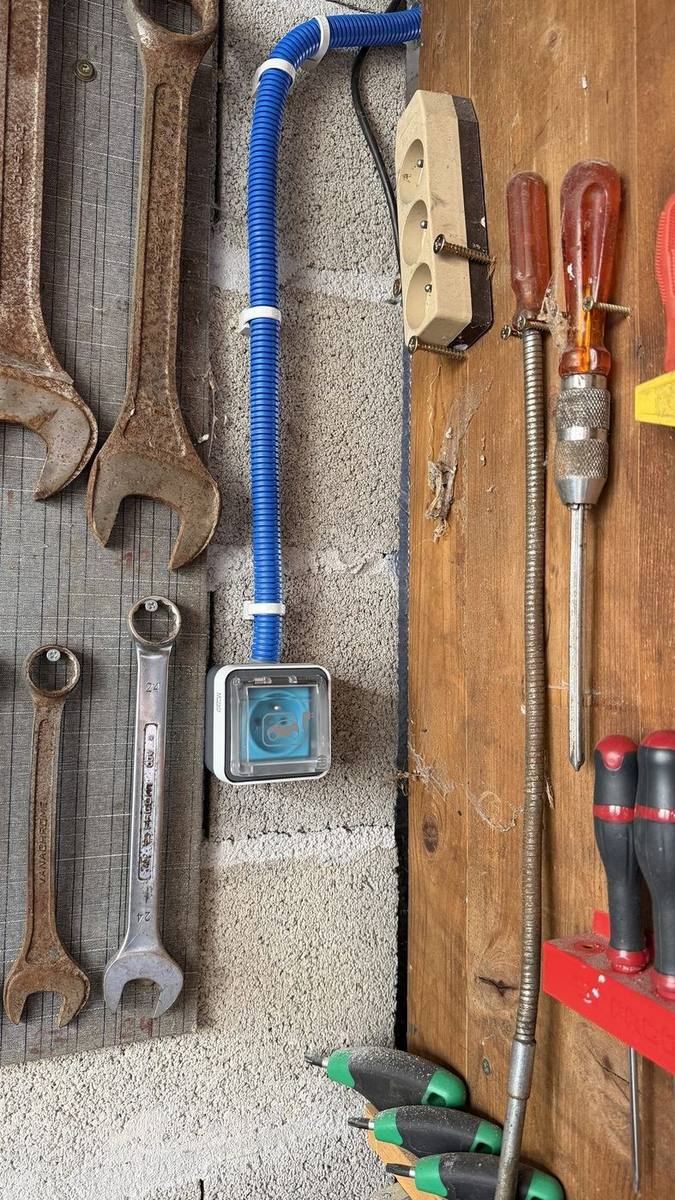 Vue plus large du mur de garage : la gaine ICTA bleue descend le long du parpaing jusqu'à la prise renforcée, entre le panneau à outils et un bloc de prises existant